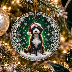 May include: A round ceramic Christmas ornament featuring a black and white Bernedoodle dog wearing a red Santa hat with the name Stella printed on the brim. The dog sits against a dark green background with white snowflakes and silver filigree.
