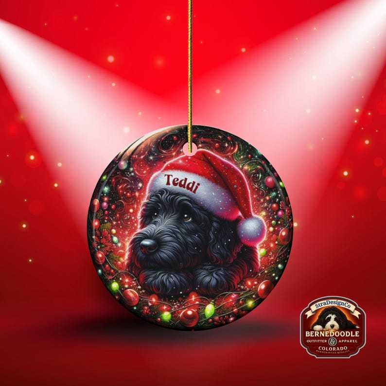 May include: A round Christmas ornament featuring a black dog wearing a Santa hat with the name "Teddi". The ornament is decorated with festive lights and ornaments against a red background. A gold string is attached for hanging.