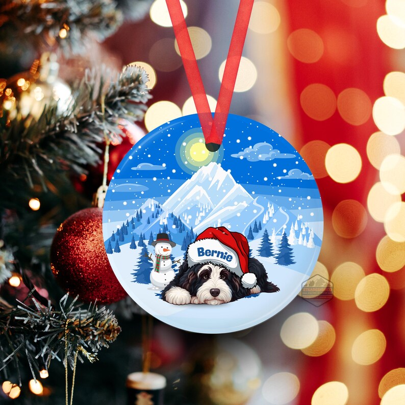 May include: Round Christmas ornament with a winter scene featuring a black and white dog wearing a Santa hat with the name "Bernie". The ornament has a red ribbon for hanging. The background shows a snowy mountain landscape with a snowman.