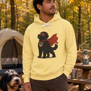 Pu&ograve; includere: Una felpa con cappuccio giallo pallido con la grafica di un cane nero che indossa un mantello e una maschera da supereroe rossi. Il cane ha un emblema a forma di cuore sul petto con strisce rosse, bianche e blu. La felpa ha lacci bianchi.