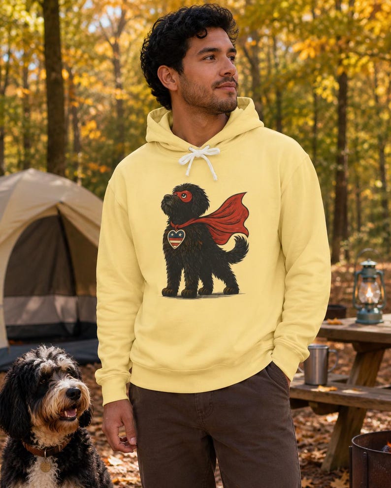 Pu&ograve; includere: Una felpa con cappuccio giallo pallido con la grafica di un cane nero che indossa un mantello e una maschera da supereroe rossi. Il cane ha un emblema a forma di cuore sul petto con strisce rosse, bianche e blu. La felpa ha lacci bianchi.