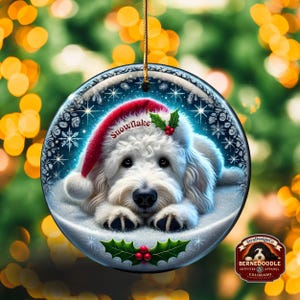May include: A round Christmas ornament featuring a white dog wearing a red Santa hat with the word "Snowflake". The dog is lying in a snowy scene with snowflakes and holly. The ornament has a gold string for hanging.