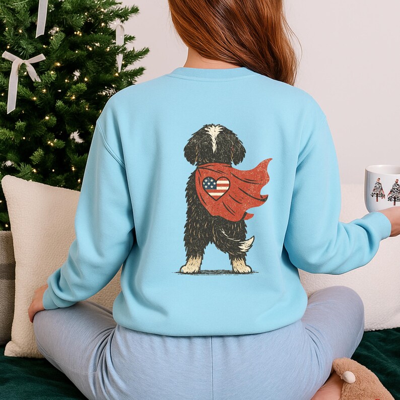 May include: Light blue sweatshirt featuring a graphic of a black and white dog wearing a red cape with a heart-shaped American flag design. The dog is standing, and the sweatshirt has long sleeves.