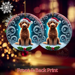 A curly golden-brown goldendoodle sits centered on a snowy ceramic ornament, wearing a Santa hat that reads “Harper.” Blue luminous sparkles, swirling winter feathers, and holly berries add a festive design touch. The ornament hangs in front of warmly lit tree lights, making it a charming holiday piece for pet enthusiasts.