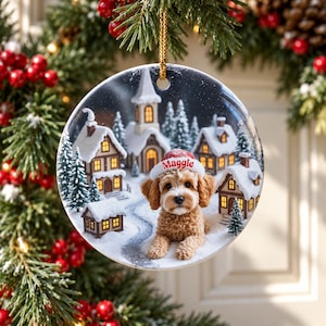 May include: A round ceramic Christmas ornament featuring a fluffy light brown dog wearing a white and red Santa hat with the name Maggie printed in red. The background shows a snowy village scene with lit houses and pine trees.