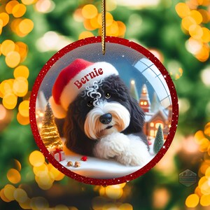 K&ouml;nnte beinhalten: Runder Weihnachtsschmuck mit einem schwarz-wei&szlig;en Hund, der einen Weihnachtsmannhut mit dem Namen "Bernie" tr&auml;gt. Der Schmuck hat einen roten Glitzerrahmen und eine goldene Schnur zum Aufh&auml;ngen. Der Hintergrund zeigt eine Winterszene mit einem Weihnachtsbaum und einem Haus.