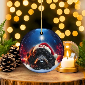 May include: A round Christmas ornament featuring a black dog wearing a Santa hat, with the name "Stella" on the hat. The ornament has a blue background with a star and snow. A gold string is attached for hanging.