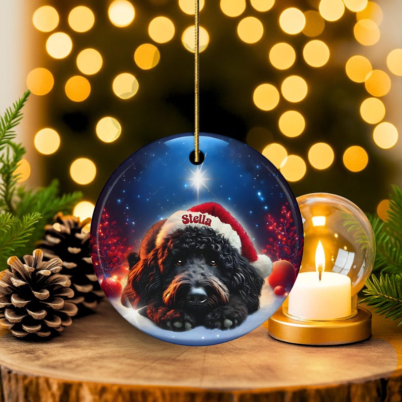 May include: A round Christmas ornament featuring a black dog wearing a Santa hat, with the name "Stella" on the hat. The ornament has a blue background with a star and snow. A gold string is attached for hanging.