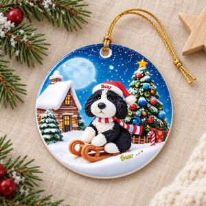A ceramic ornament featuring a black and white dog wearing a Santa hat and scarf, sitting on a sled and surrounded by a snowy village scene with a Christmas tree and a full moon.