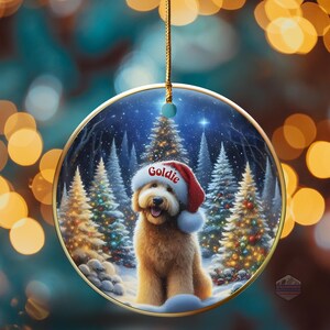 May include: A round Christmas ornament featuring a golden retriever wearing a Santa hat with the name "Goldie". The ornament has a winter scene with snow-covered trees and a gold trim. A gold string is attached for hanging.