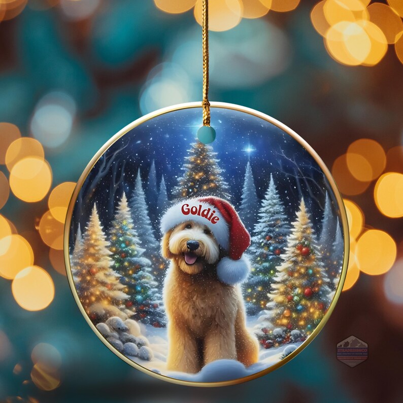 May include: A round Christmas ornament featuring a golden retriever wearing a Santa hat with the name "Goldie". The ornament has a winter scene with snow-covered trees and a gold trim. A gold string is attached for hanging.