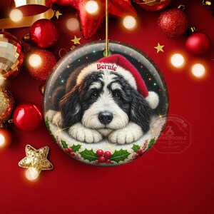 May include: A round Christmas ornament featuring a Bernedoodle dog wearing a Santa hat. The dog's name, "Bernie," is printed on the hat. The ornament is surrounded by red and gold Christmas decorations.