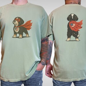 May include: Sage green t-shirts featuring a cartoon dog superhero with a red cape and a heart-shaped emblem. The front of the shirt shows the dog facing forward, while the back shows the dog's rear view. The dog has a mask and the emblem has a patriotic design.