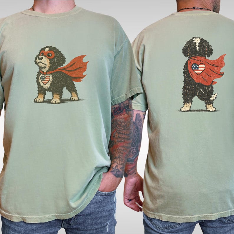 May include: Sage green t-shirts featuring a cartoon dog superhero with a red cape and a heart-shaped emblem. The front of the shirt shows the dog facing forward, while the back shows the dog's rear view. The dog has a mask and the emblem has a patriotic design.