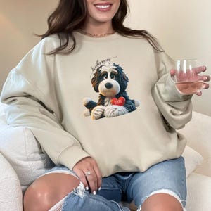 A woman wearing a Bernedoodle sweatshirt with cute design on the front and the back.  The front features a bernedoodle dog in claymation design holding a heart with his name, across the top of his head the words, "Doodle Mom".