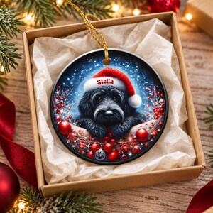 May include: A round ceramic Christmas ornament featuring a black dog wearing a red Santa hat with the name Stella printed on the brim. The dog is surrounded by red berries, red ornaments, and a snowy blue background with a gold hanging cord.