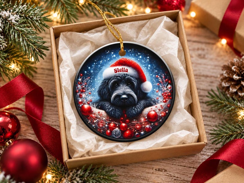 May include: A round ceramic Christmas ornament featuring a black dog wearing a red Santa hat with the name Stella printed on the brim. The dog is surrounded by red berries, red ornaments, and a snowy blue background with a gold hanging cord.