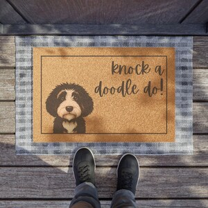 May include: A rectangular doormat with a brown center and a blue and white plaid border. The mat features a black and white doodle dog illustration and the phrase "knock a doodle do!" in black script. The mat is placed on a wooden porch.