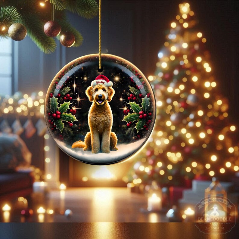 May include: A round Christmas ornament featuring a golden doodle dog wearing a Santa hat with the name "Goldie". The ornament is surrounded by holly and berries, with a starry background. A gold string is attached for hanging.