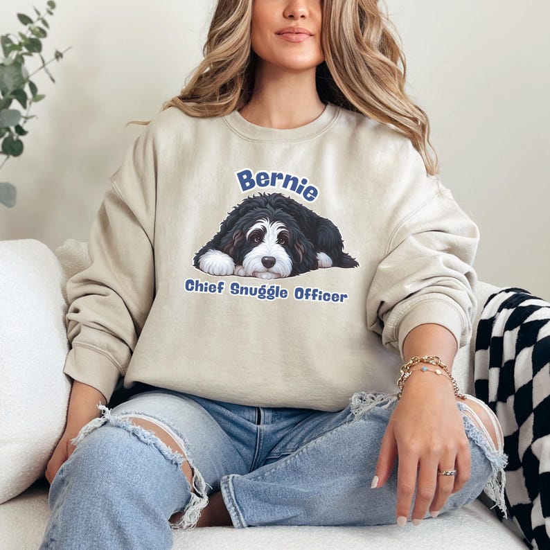Puede incluir: Sudadera beige con un dise&ntilde;o gr&aacute;fico de un perro de monta&ntilde;a bern&eacute;s. El perro es negro, blanco y marr&oacute;n, con el nombre "Bernie" arriba y "Chief Snuggle Officer" abajo. La sudadera es de color neutro.