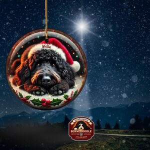 May include: A round Christmas ornament featuring a black dog wearing a Santa hat. The dog's name, "Stella," is printed on the hat. The ornament is decorated with holly and berries and hangs against a snowy mountain backdrop.