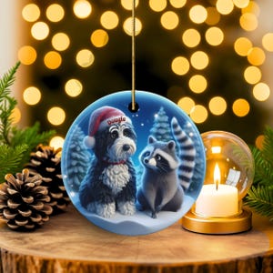A 3-inch double-sided ceramic Christmas ornament featuring an adorable claymation-style Bernedoodle wearing a red Santa hat personalized  sitting beside a cute raccoon under a snowy winter night sky. The ornament has crisp detailing, soft blue tones, and a warm Christmas glow in the blurred background. Perfect for Bernedoodle lovers, handmade Christmas gift shoppers. High-quality ceramic with vibrant colors, ideal for tree decorating, pet memorial ornaments, or festive seasonal displays.