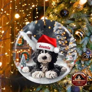 May include: A round Christmas ornament featuring a black and white Bernedoodle dog wearing a red Santa hat with the name "Zoomie". The ornament has a winter scene background with a snowy house and trees. The ornament is hanging from a gold string.