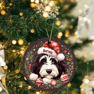 May include: Round Christmas ornament featuring a black and white dog wearing a Santa hat with the name "Bernie". The ornament is decorated with red and white ornaments, snowflakes, and a wreath of greenery and flowers. A gold string is attached for hanging.