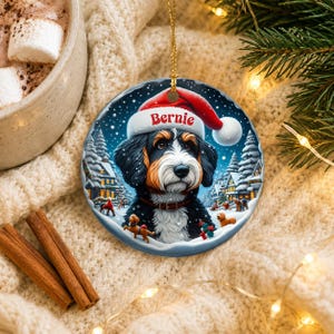 A Christmas ornament featuring a Bernese mountain dog wearing a Santa hat, surrounded by a snowy winter scene with houses, trees, and people.