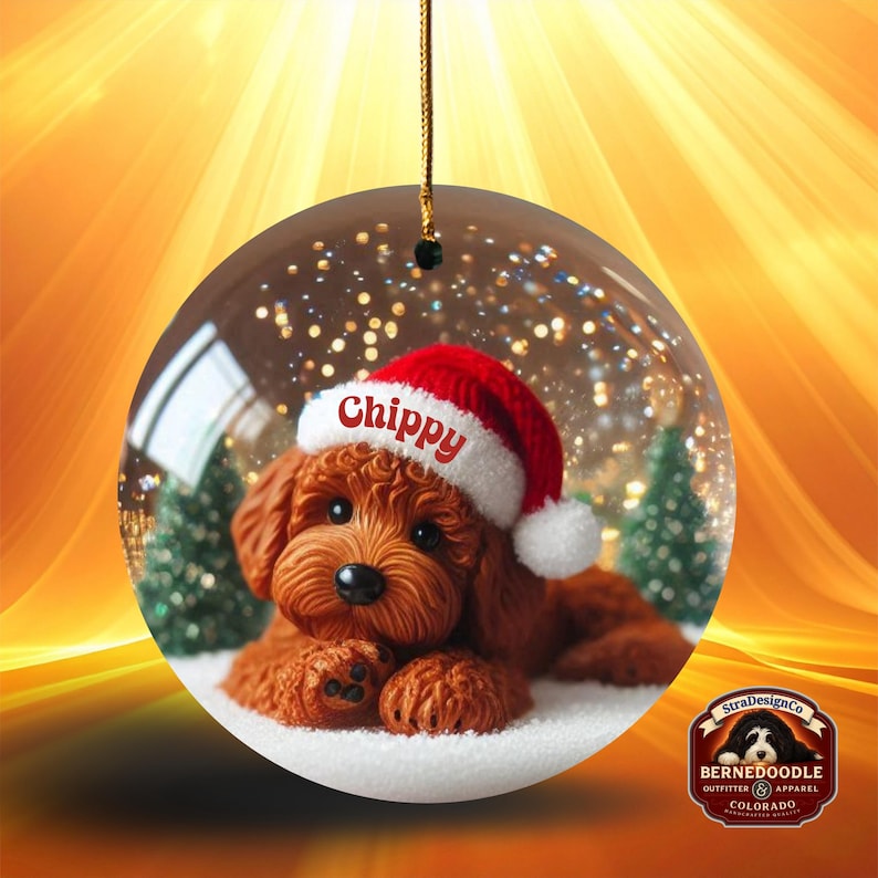 May include: A circular Christmas decoration showing a brown dog wearing a red Santa hat with the name "Chippy". The dog is inside a clear glass ball with a snowy base and small green trees. The ornament hangs from a gold string.