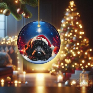 May include: A round Christmas ornament featuring a black dog wearing a Santa hat with the name "Stella". The ornament has a blue background with a star and snow. The ornament is hanging from a gold string, with a Christmas tree in the background.