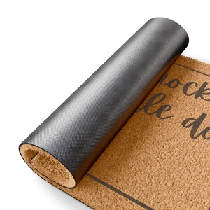 May include: A rolled-up brown doormat with black text that reads "knock le do". The mat has a dark gray, textured backing. The doormat is made of a natural fiber material.