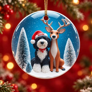 May include: A round ceramic Christmas ornament featuring a black and white dog wearing a red Santa hat with the name Bernie printed on it, sitting next to a brown deer in a snowy forest with pine trees under a blue sky.