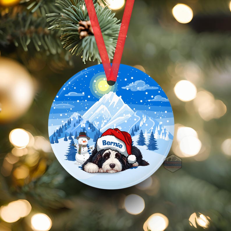 May include: Round Christmas ornament featuring a Bernese Mountain Dog wearing a Santa hat, with the name "Bernie" on the hat. The ornament has a winter scene with snow, mountains, a snowman, and a full moon. A red ribbon is attached for hanging.