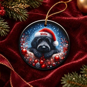 May include: A round ceramic Christmas ornament featuring a black dog wearing a red Santa hat with the name Stella printed on the brim. The illustration includes red berries and ornaments against a snowy blue background with a gold hanging cord.