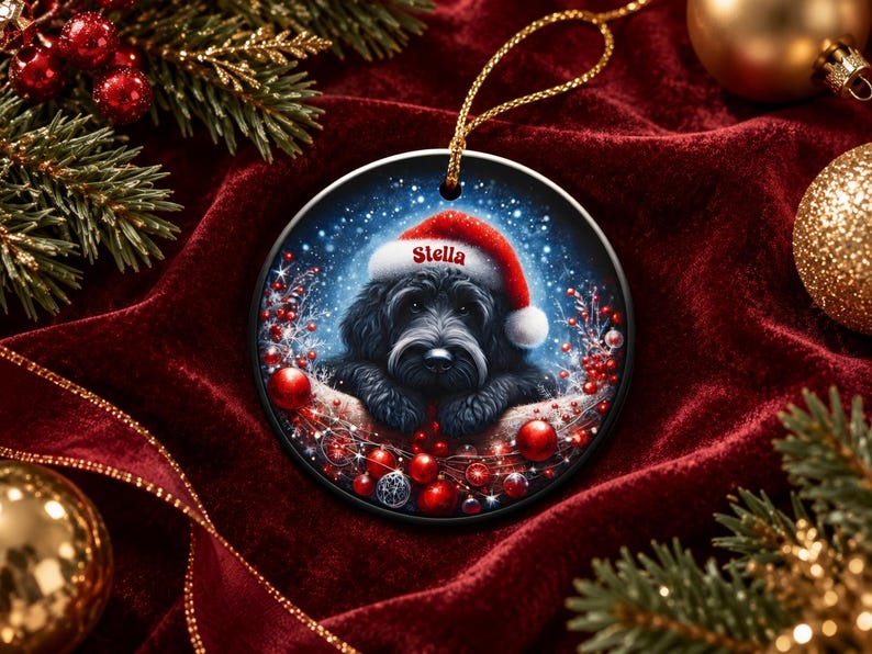 May include: A round ceramic Christmas ornament featuring a black dog wearing a red Santa hat with the name Stella printed on the brim. The illustration includes red berries and ornaments against a snowy blue background with a gold hanging cord.