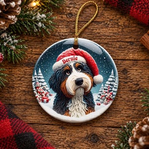 May include: A round ceramic Christmas ornament featuring a Bernese Mountain Dog wearing a red Santa hat with the name Bernie printed on the brim. The dog is set against a snowy winter landscape with pine trees and small snowmen.