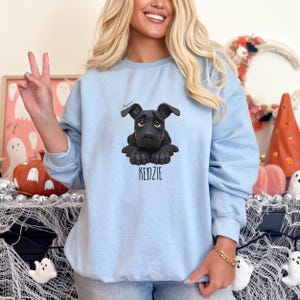 Puede incluir: Sudadera azul claro con una ilustraci&oacute;n de un perro negro y el nombre "KENZIE" impreso debajo. El perro tiene una expresi&oacute;n triste y est&aacute; acostado. La sudadera es de material suave.