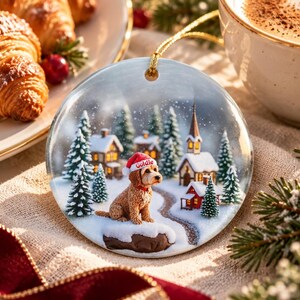 May include: A round ceramic Christmas ornament featuring a golden-brown dog wearing a red Santa hat with the name Goldie printed on it. The scene depicts a snowy village with pine trees, small houses, and a church under a light snowfall.