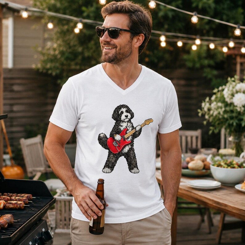 May include: A white V-neck t-shirt featuring a graphic illustration of a black and white dog playing a red electric guitar. The dog is depicted in a standing position, holding the guitar with its front paws while wearing a small collar.