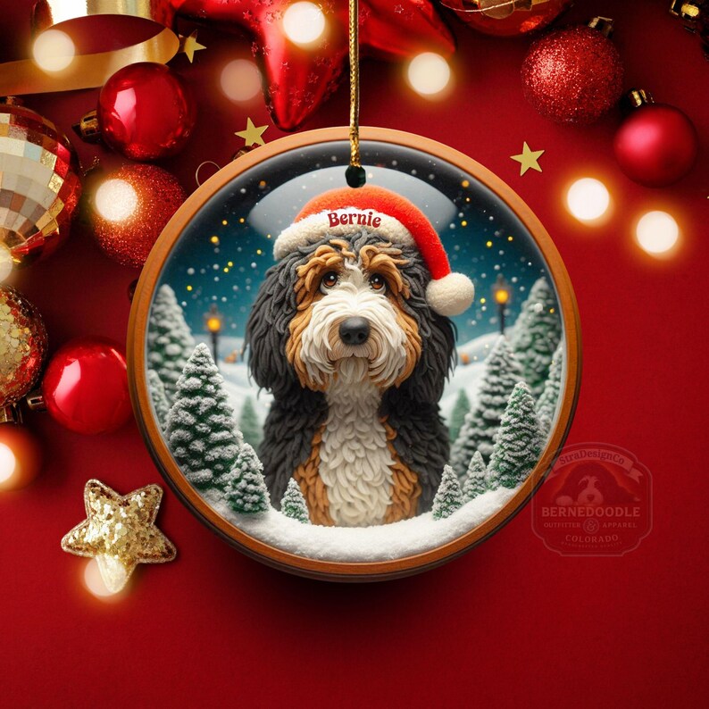 Op de afbeelding: Een ronde kerstversiering met een Bernedoodle-hond met een kerstmuts met de naam "Bernie". De versiering heeft een winterse achtergrond met besneeuwde bomen en een besneeuwde grond. De versiering hangt tegen een rode achtergrond met kerstversieringen.