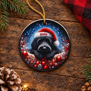 May include: A round ceramic Christmas ornament featuring a black dog wearing a red Santa hat with the name Stella printed on the brim. The dog is surrounded by red ornaments, berries, and a snowy background, hanging from a gold string.
