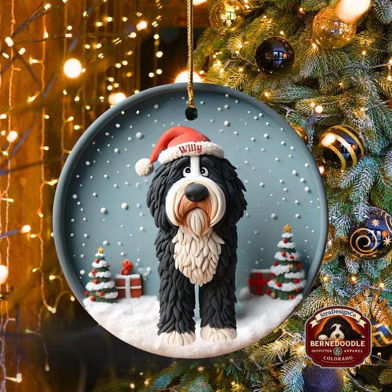 May include: A round Christmas ornament featuring a black and white Bernedoodle dog wearing a Santa hat with the name "Willy". The ornament has a snowy scene with Christmas trees and gifts. The ornament is hanging from a gold string.