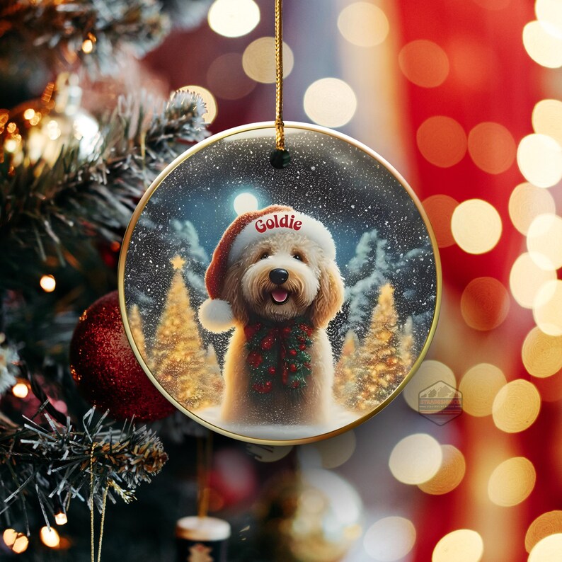 May include: A round Christmas ornament featuring a smiling Goldendoodle wearing a Santa hat with the name "Goldie" and a festive scarf. The background shows a snowy forest scene with Christmas trees and a full moon. The ornament has a gold trim.