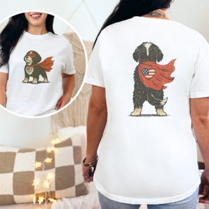 May include: White t-shirt featuring a cartoon dog dressed as a superhero. The dog has a red cape and mask, with a heart-shaped emblem on its chest. The back of the shirt also displays the dog with the cape.