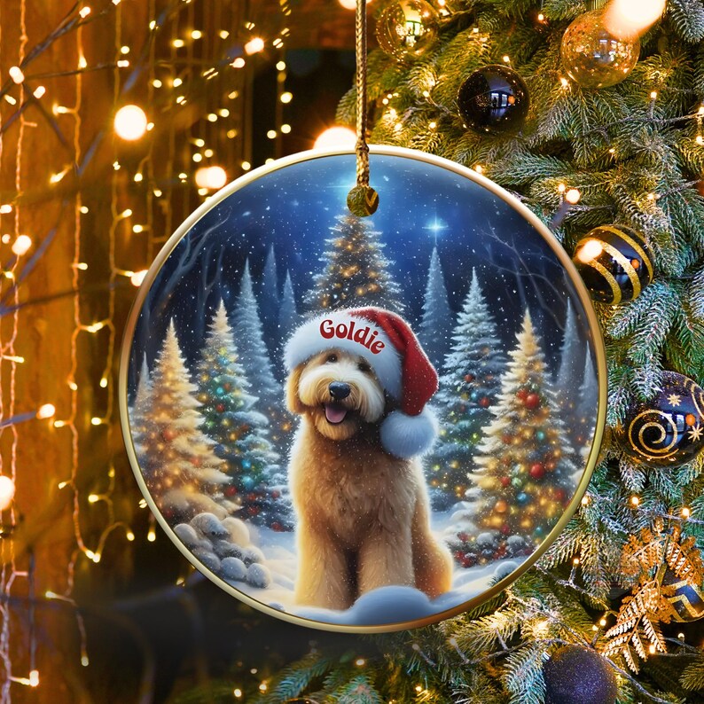 May include: Round Christmas ornament featuring a golden doodle dog wearing a Santa hat with the name "Goldie" on it. The background shows a snowy forest scene with Christmas trees. The ornament is hanging from a gold string.