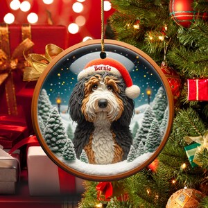 Op de afbeelding: Ronde kerstversiering met een Bernedoodle-hond met een kerstmuts met de naam "Bernie". De versiering heeft een houten frame en een winterse achtergrond met besneeuwde bomen en een sterrenhemel. Een feestelijke vakantieversiering.