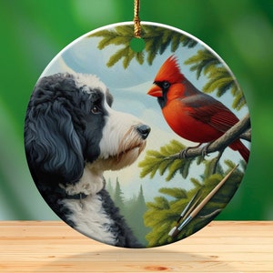 May include: Round ceramic ornament featuring a black and white dog gazing at a vibrant red cardinal perched on a branch. The artwork includes green pine branches and a forest backdrop, with paintbrushes visible. A gold string is attached for hanging.