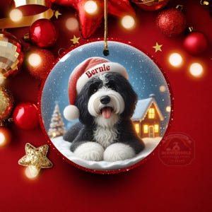 May include: A round Christmas ornament featuring a black and white Bernedoodle dog wearing a Santa hat with the name "Bernie". The ornament has a snowy winter scene with a house and Christmas tree. The ornament is surrounded by red Christmas decorations.