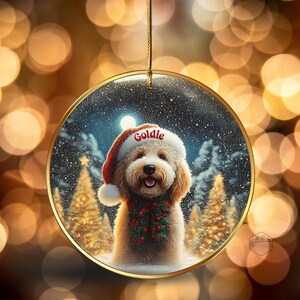 May include: Round Christmas ornament featuring a fluffy dog wearing a Santa hat with the name "Goldie" and a festive garland. The background shows a snowy winter scene with Christmas trees and a full moon. The ornament has a gold-colored border and a gold string for hanging.